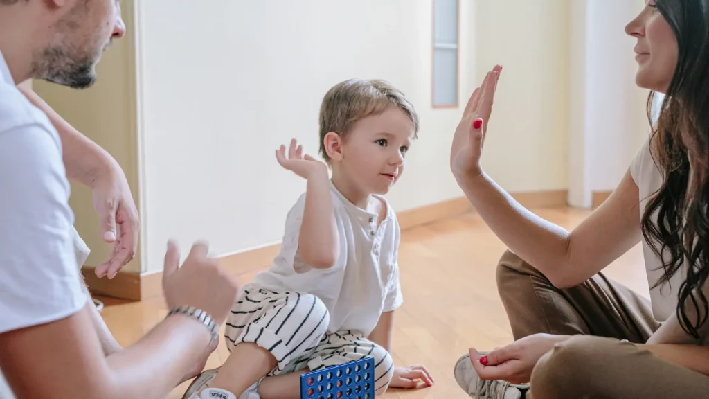 Young child sitting on the floor, smiling and giving a high-five to an adult during a game.