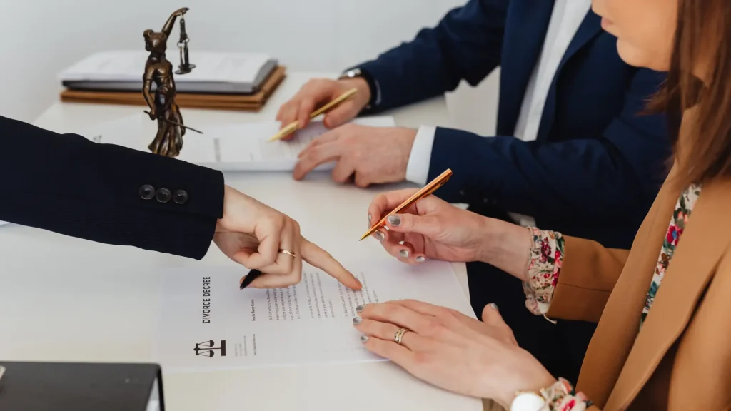 Multiple people in a business meeting are signing legal documents, with one person pointing to a section on the paper